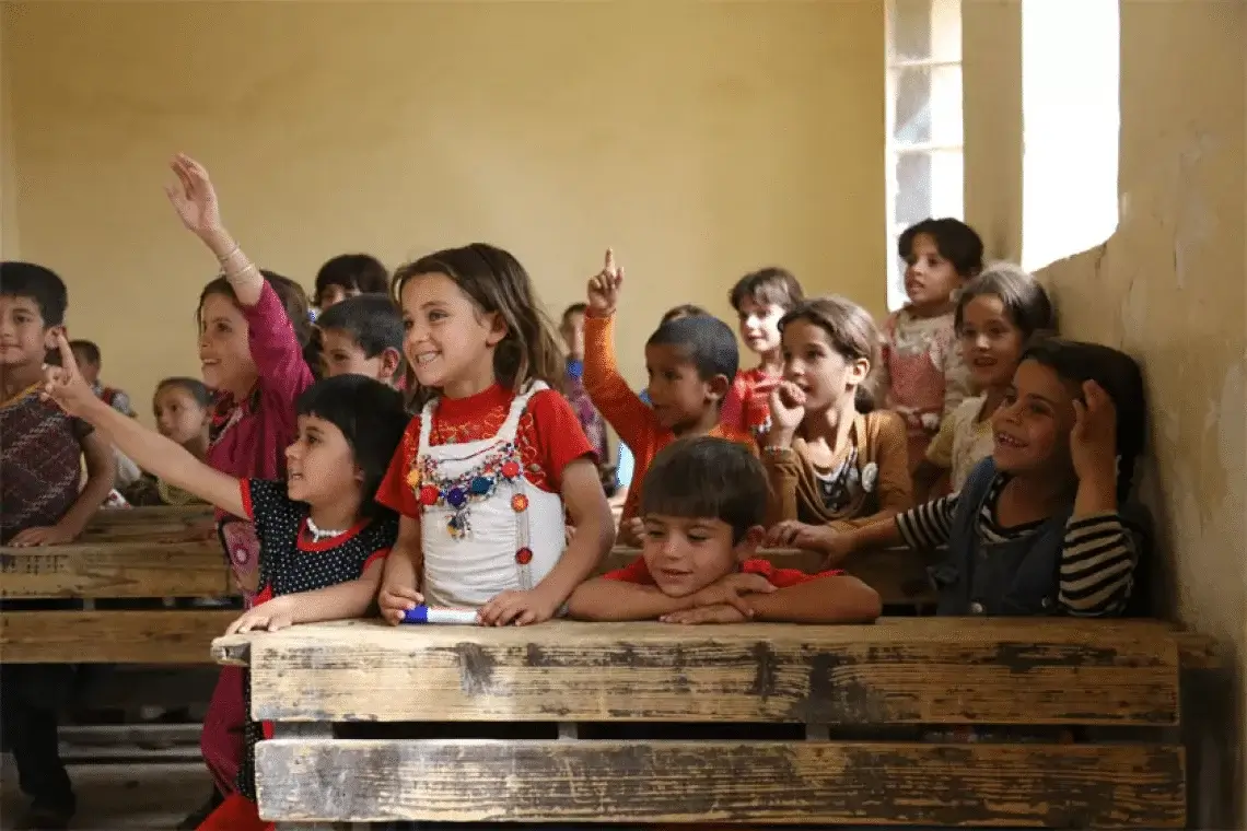 A group of young students in a rural classroom raising their hands to answer questions, sitting at wooden desks with bright smiles and eager expressions