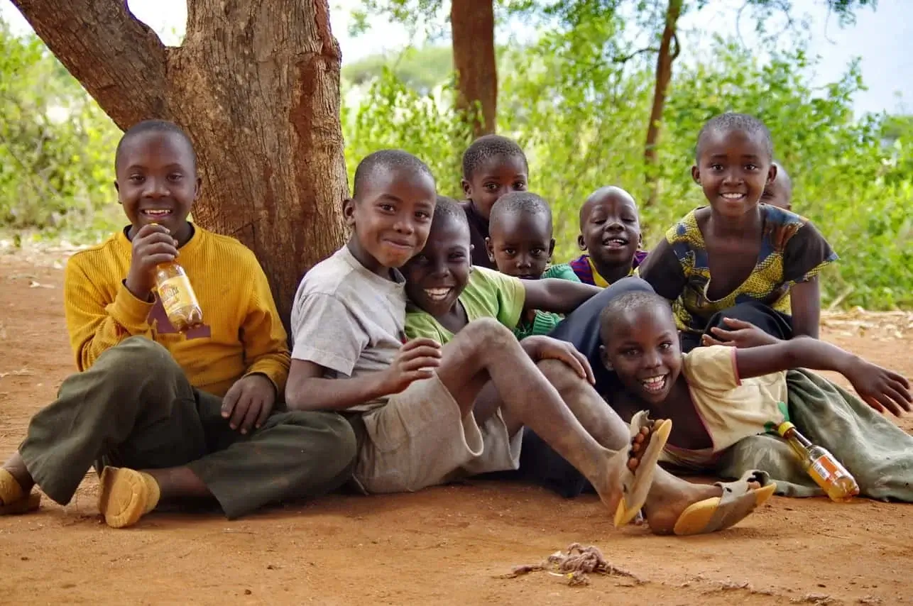 A group of smiling African boys sitting under a tree on red soil, laughing and enjoying each other's company in a rural outdoor setting