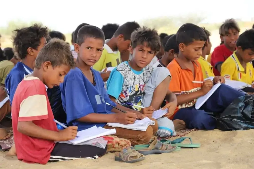 A group of young boys sitting on the ground outdoors, writing in notebooks with pens — a scene of education in a resource-limited environment
